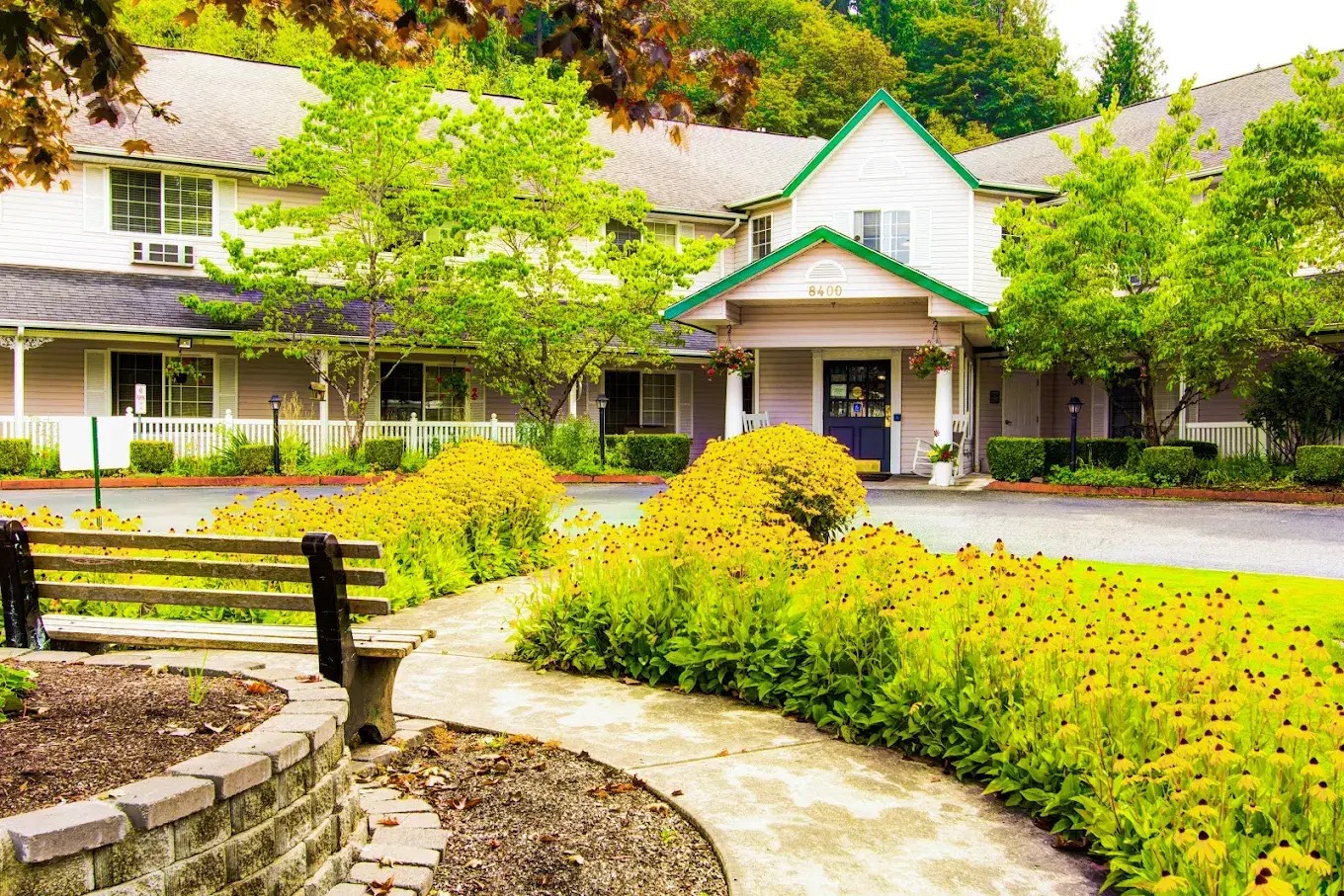 Front entrance to Cascade Valley Senior Living in Arlington, Washington, a cream two storey assisted living and memory care building with a green gabled porch marked 8400, a white picket fence and black-eyed Susans blooming along the garden path
