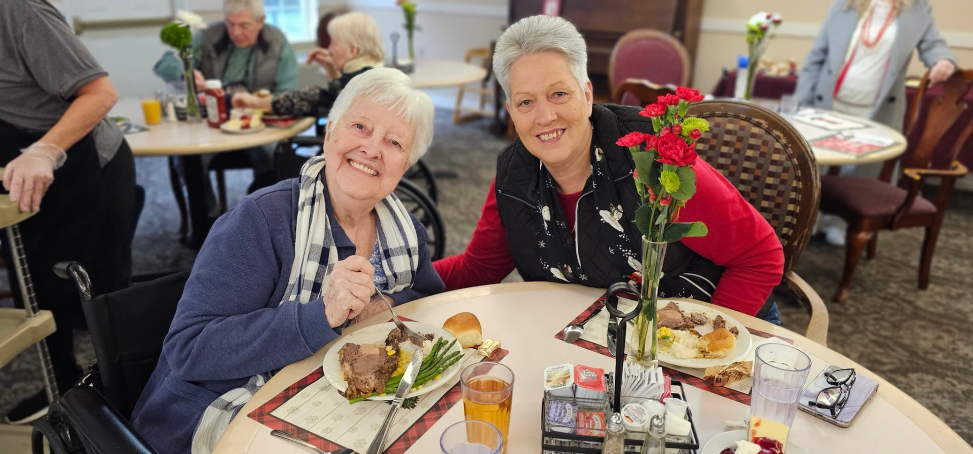 Two residents sharing lunch at a round table in the Cascade Valley Senior Living dining room in Arlington, Washington, with fresh flowers and a plated meal